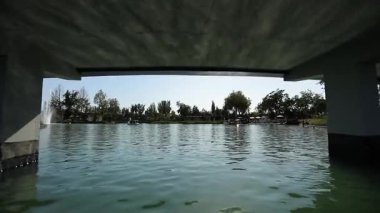 Ağaçlarla çevrili bir parktaki göl suyunda bir dron. Güverte köprüsünün manzarası. Bulutsuz bir yaz günü. Hava Görünümü