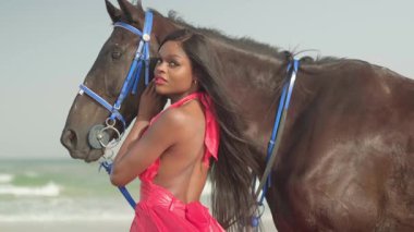 Kırmızı elbiseli genç esmer bir kadın okyanus kıyısında bir atın yanında duruyor. Yavaş çekim.