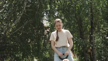 Şortlu genç bir anne, küçük oğlunu oyuncak bir arabaya bindiriyor. Park, güneşli bir gün. Yavaş Hareket.