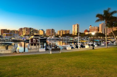 Sarasota 'nın rıhtımının büyüleyici manzarası marina boyunca kenetlenmiş sayısız teknenin alacakaranlık gökyüzüne karşı aydınlatılmış canlı şehir silueti..