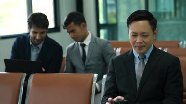 Asyalı işadamı, havaalanındaki koltukta otururken akıllı telefondaki verileri kontrol ediyor. İki genç iş adamı arkada oturmuş laptoplar ve tabletler aracılığıyla bilgi alışverişinde bulunuyor..