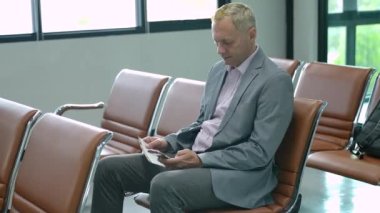 İş adamı uçuş kartlarını kontrol ediyor. İnsanlar havaalanı sandalyelerinde oturmuş uçağa binmeyi bekliyor..