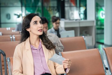 İş kadını uçuş kartlarını kontrol ediyor. Havaalanı sandalyelerinde oturan insanlar uçağa binmek için bekliyor..