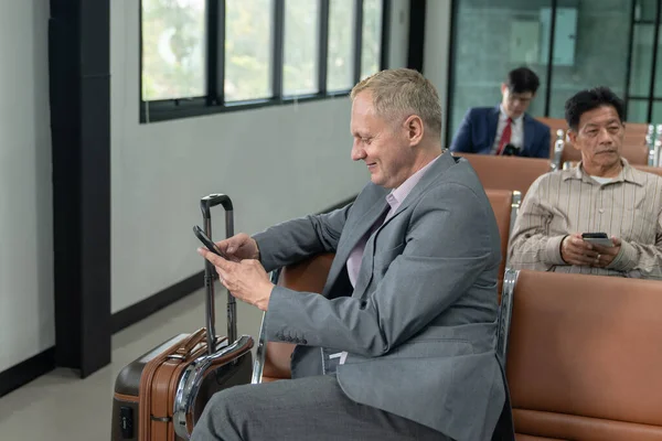 Businessman sitting on chair meeting remotely via mobile phone. Man smiling happily while playing social media. Businessman waiting to board the plane inside the airport. Side view of man.