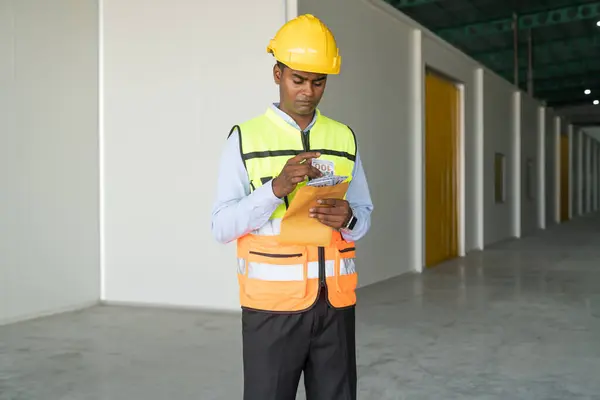 Indian male engineer in safety helmet and reflective vest holding prize money envelope in warehouse.