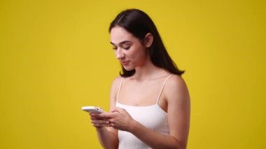Sarı arka planda izole edilmiş telefonundan bir şey izleyen hoş bir kadının 4K videosu. Teknoloji kavramı.