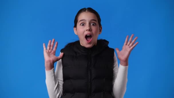 青を背景に大きな目でカメラを見て驚きの手を挙げている女の子の4K動画 感情の概念 — ストック動画