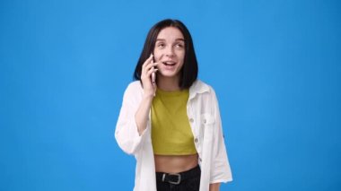 Mavi arka planda telefonla konuşan hoş bir kızın 4K videosu. Duyguların kavramı.