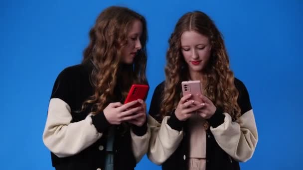4K个双胞胎女孩在蓝色背景下使用手机的视频 情绪的概念 — 图库视频影像