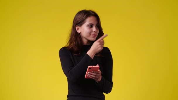 4K慢动作视频 一个女孩拿着电话指着右边 用大拇指在黄色背景上显示 情绪的概念 — 图库视频影像