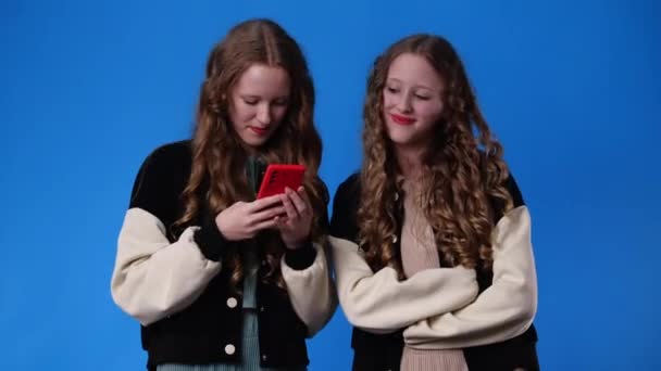 青い背景の上に携帯電話を使って双子の女の子の4Kのスローモーションビデオ 感情の概念 — ストック動画