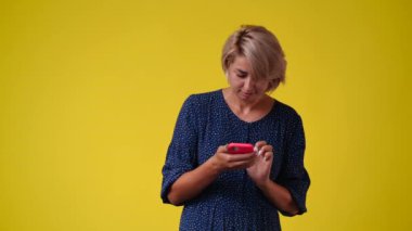 4k video of one woman typing text on the phone and looking around over yellow background. Concept of emotions.
