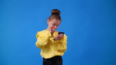 4k video of one young girl who scrolling while looking at the phone and smiling over blue background. Concept of emotion.