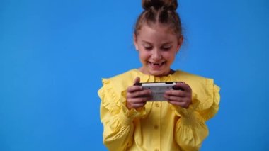 4k video of one young girl is playing on the phone and smiling over blue background. Concept of emotions.