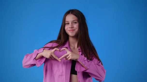 Parmaklarıyla Kalbinin Işaretini Gösteren Mavi Arka Planda Gülümseyen Bir Kızın — Stok video