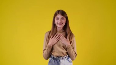 4k slow motion video of one girl who smiles sweetly and puts his hands to his cheeks over yellow background. Concept of emotions.