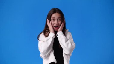 4k video of girl raising hands in surprise looking at camera with big eyes on blue background. Concept of countdown.