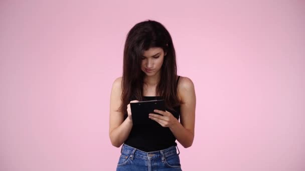 ピンクの背景の上にタブレットでペンで何かを考え メモする1人の女の子の4Kビデオ 感情の概念 — ストック動画