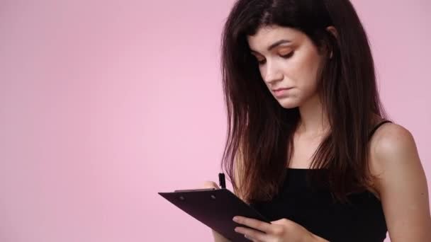 ピンクの背景の上にタブレットでペンで何かを考え メモする1人の女の子の4Kのクローズアップビデオ 感情の概念 — ストック動画