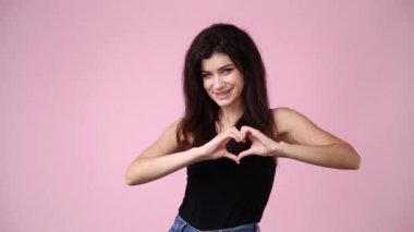 Pembe arka planda elleri ile kalp şekli oluşturan bir kızın 4K videosu. Aşk duyguları kavramı.