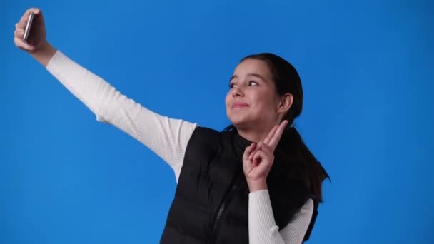 青い背景の上に自分の携帯電話でセルフィーを取っている1人の女の子の4Kビデオ 感情の概念 — ストック動画