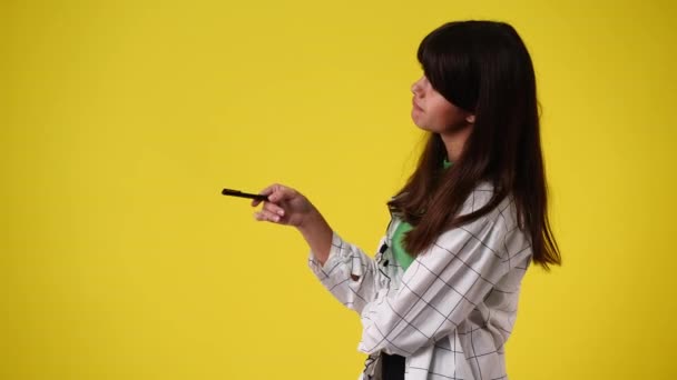 黄色の背景の上にペンを手に何かを考えて計画している1人の少女の4Kビデオ 感情の概念 — ストック動画