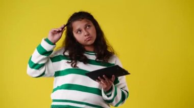 Bir kızın 4k videosu, sarı arkaplandaki tablete kalemle ilgili bir şey düşünüyor ve not alıyor. Duyguların kavramı.