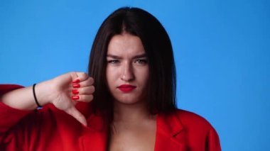 Negatif yüz ifadesi olan bir kadının mavi arka planda baş parmağını aşağı gösteren 4k yavaş çekim videosu. Duyguların kavramı.