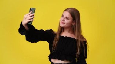 Telefonunda selfie çeken ve sarı arka planda barış işareti gösteren bir kızın 4K videosu. Duyguların kavramı.