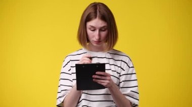 Bir kızın 4k videosu, sarı arkaplandaki tablete kalemle ilgili bir şey düşünüyor ve not alıyor. Duyguların kavramı.