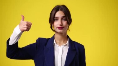 Yüzünde belirsiz bir ifade olan bir kızın sarı arka planda baş parmağı yukarı ve baş parmakları aşağı gösteren 4K videosu. Duyguların kavramı.