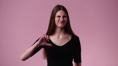 Yüzünde negatif ifade olan bir kızın pembe arka planda baş parmağını aşağı gösteren 4K videosu. Duyguların kavramı.