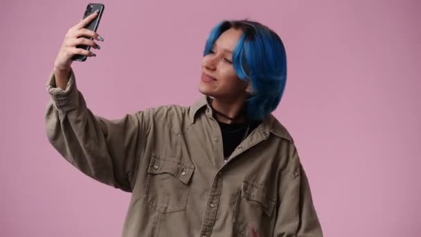 4K视频 一个女孩在他的手机上自拍 并在粉色背景上显示和平标志 情绪的概念 — 图库视频影像