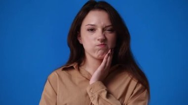 Mavi arka planda diş ağrısından muzdarip bir kızın 4K videosu. Duyguların kavramı.