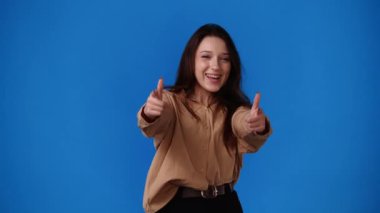 Mavi arka planda baş parmak gösteren ve gülümseyen bir kızın 4K videosu. Duyguların kavramı.