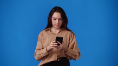 Mavi arka planda telefonu kullanan güzel bir kızın 4K videosu. Duyguların kavramı.