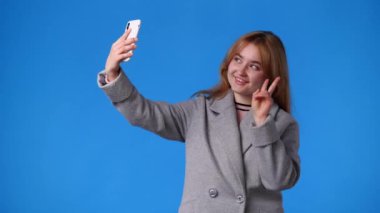 Mavi arka planda telefonundan selfie çeken bir kızın 4K videosu. Duyguların kavramı.