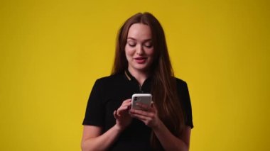 Sarı arka planda gülümseyen ve telefon kullanan bir kızın 4K videosu. Duyguların kavramı.