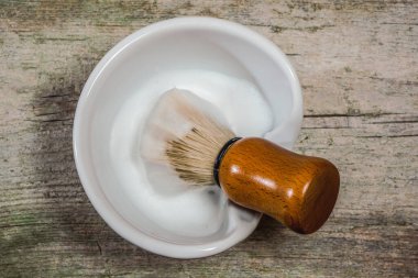 Seramik kasede traş fırçası olan sabunlu yaban domuzu makrosunun kırsal ahşap yüzeyde dinlenirken çekilmiş yatay fotoğrafı. Geleneksel ıslak tıraş malzemeleri..