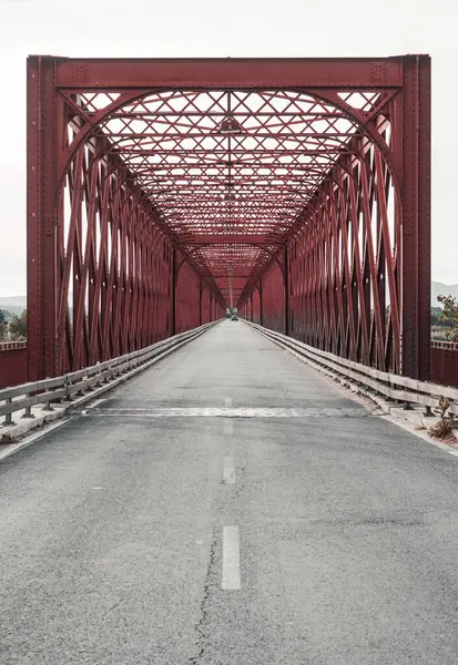 stock image A red truss bridge with a road passing under it, showcasing symmetry and a rectangular design. The bridge is made of metal and includes a walkway for pedestrians