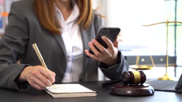 スマートフォンを閲覧してペンでメモを書く女性の部分的なビュー テーブルの上の法律ハンマー — ストック動画