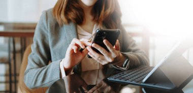 Kadın elleri dizüstü bilgisayarda yazı yazıyor ve modern ofiste akıllı telefon tutuyor.