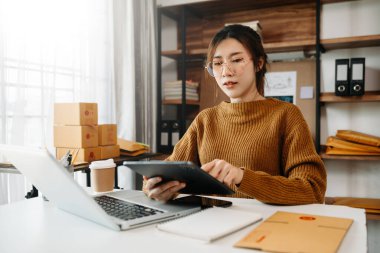 Startup small business SME, Entrepreneur owner woman using smartphone or tablet taking receive and checking online purchase shopping order to prepare pack product boxes.