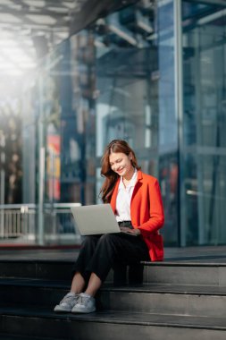 Entrepreneur holding and working on laptop while sitting at the urban street. Beautiful, businesswoman carrying laptop  and doing business outside in the city street