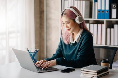 Attractive happy young Asian student studying at the college library, sitting at the desk, using a laptop computer,  and headphones having a video chat