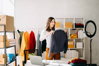 Beautiful lady blogger showing clothes in front of the camera to recording vlog video live-streaming at her shop.Online Shopping Cart notion.
