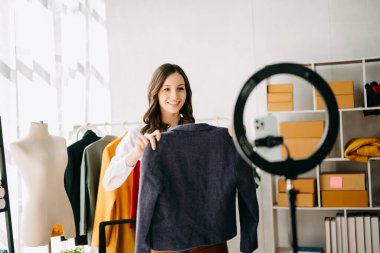 Beautiful lady blogger showing clothes in front of the camera to recording vlog video live-streaming at her shop.Online Shopping Cart notion.