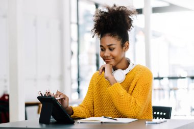 Attractive happy young African student studying at the college library, sitting at the desk, using  tablet and headphones having a video chat.