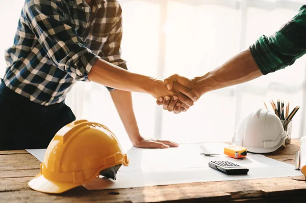 Construction team shake hands greeting start new project plan behind yellow helmet on desk in office to consults about their building project. in sunlight