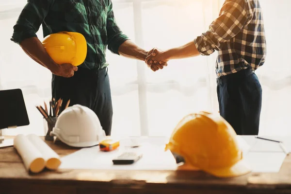 Construction team shake hands greeting start new project plan behind yellow helmet on desk in office to consults about their building project. in sunlight